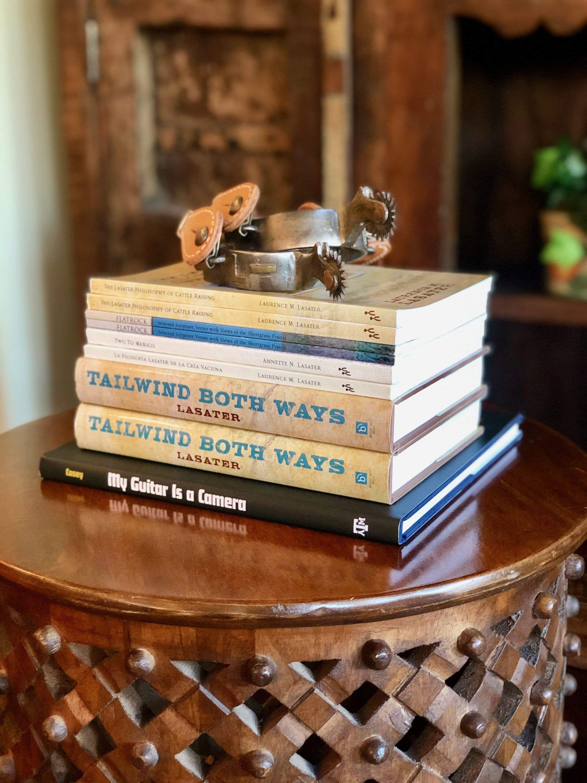 Stack of books including "Tailwind Both Ways" and "My Guitar Is a Camera" on a wooden table with a decorative design, accompanied by a vintage metal spur. The background features a rustic wooden cabinet and a green plant.
