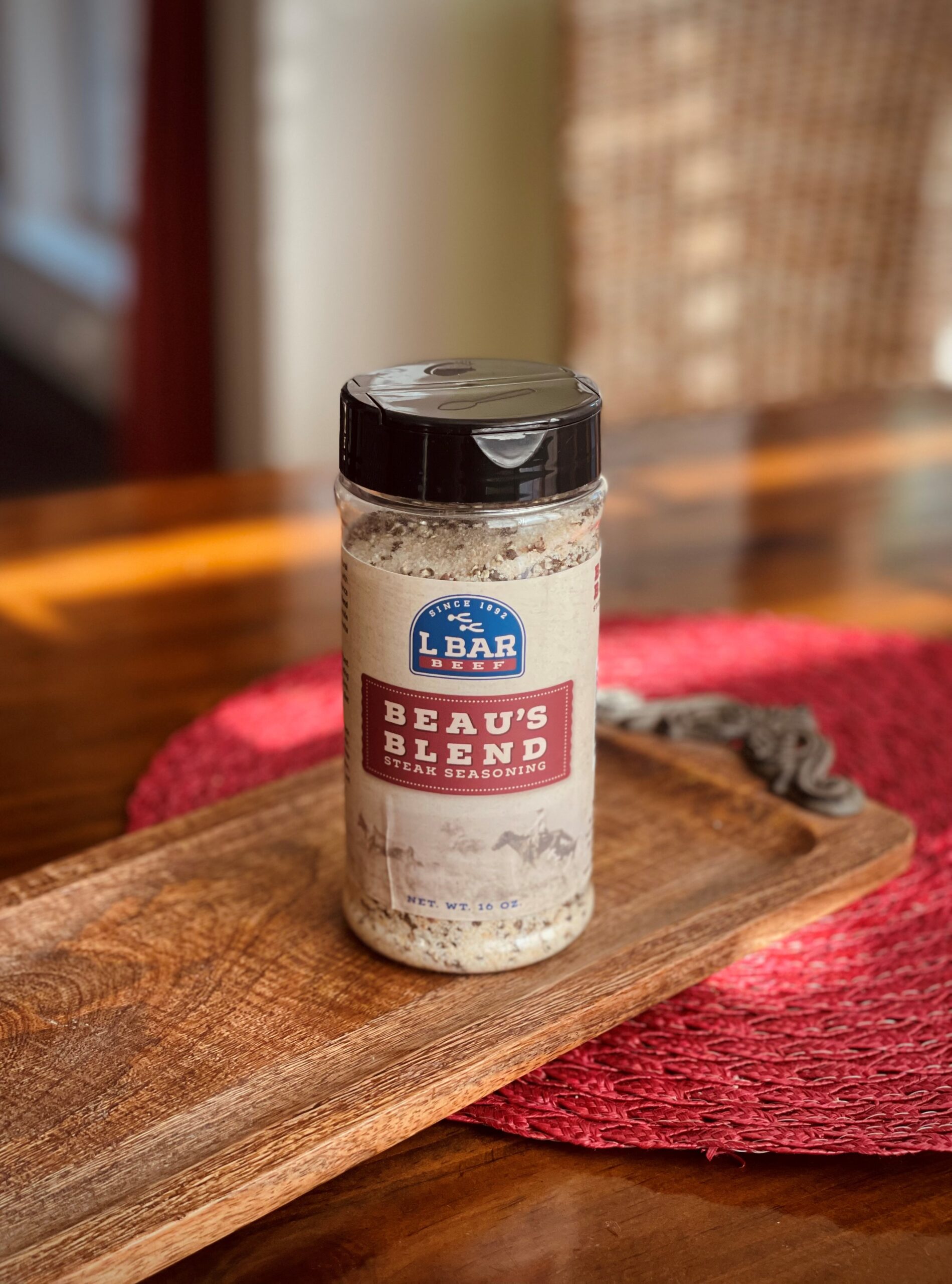 Beau's Blend steak seasoning in a clear bottle on a wooden serving tray, with a red woven placemat in the background, showcasing its 16 oz size and branding. Ideal for enhancing the flavor of grilled meats and other dishes.